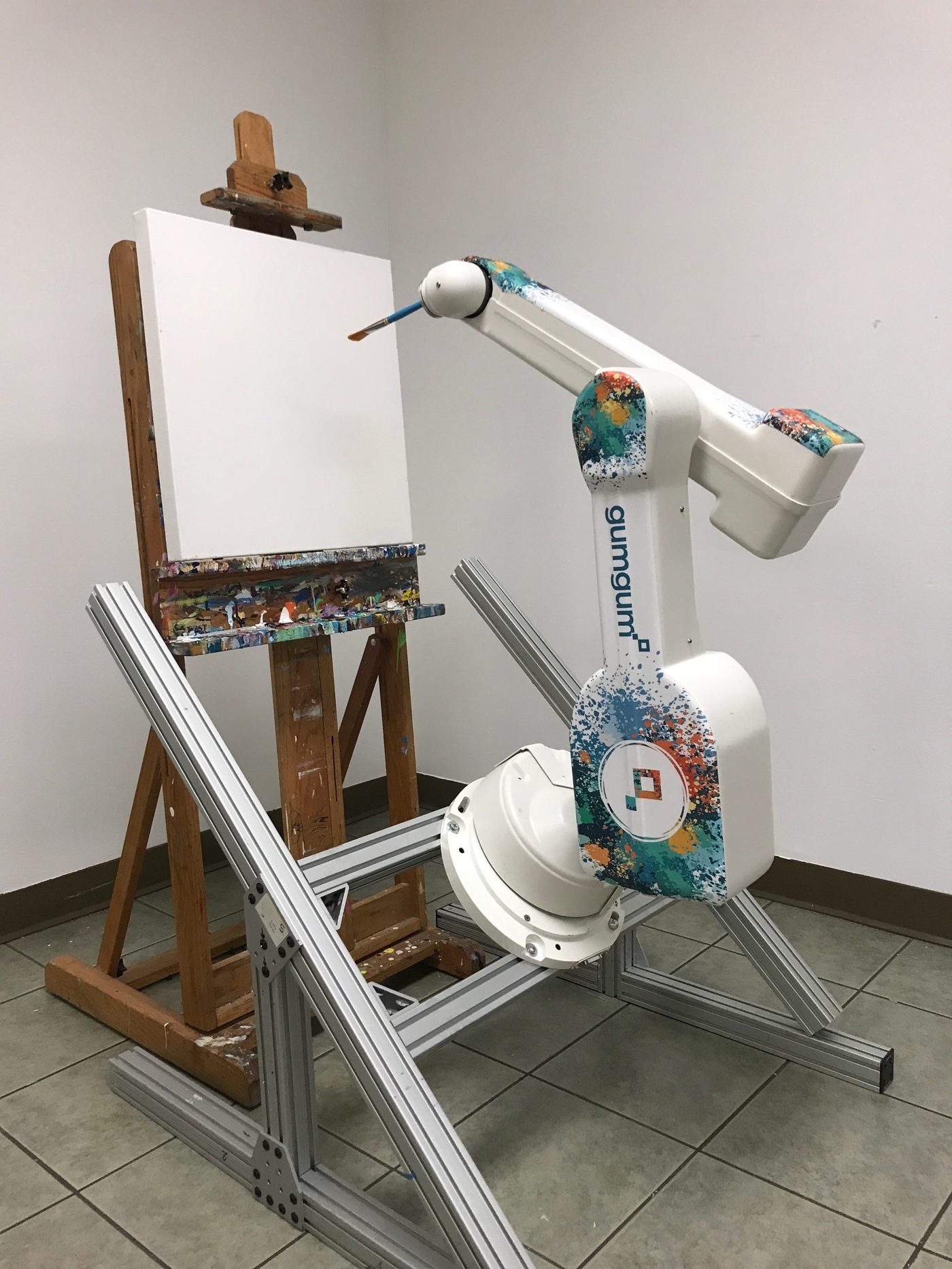 A white robot arm, wrapped in paint-splash graphics and a GumGum logo, holds a thin brush to a blank white canvas on a paint-stained wooden easel. The arm stands on an aluminium frame on a tiled floor.
