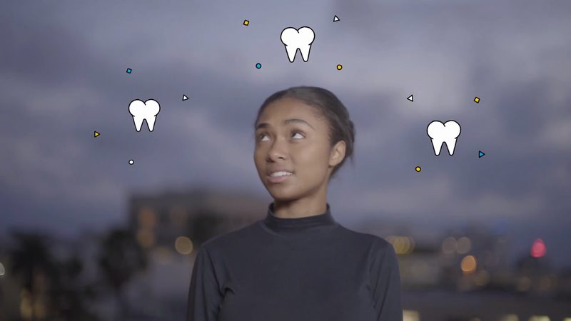 A video frame: a woman looks up at small floating tooth icons against a dusky city skyline.