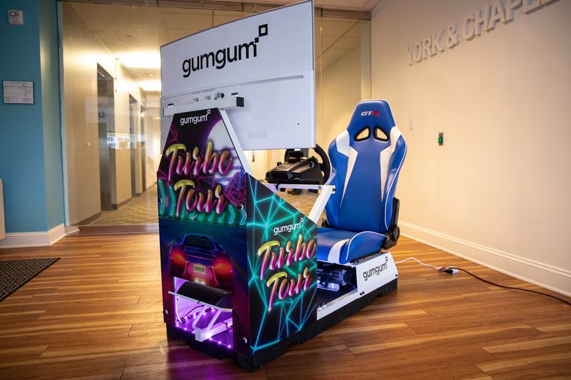 A racing-simulator arcade cabinet in an office lobby: a blue racing seat, a wheel and a large screen, the cabinet wrapped in neon Turbo Tour art and GumGum logos.
